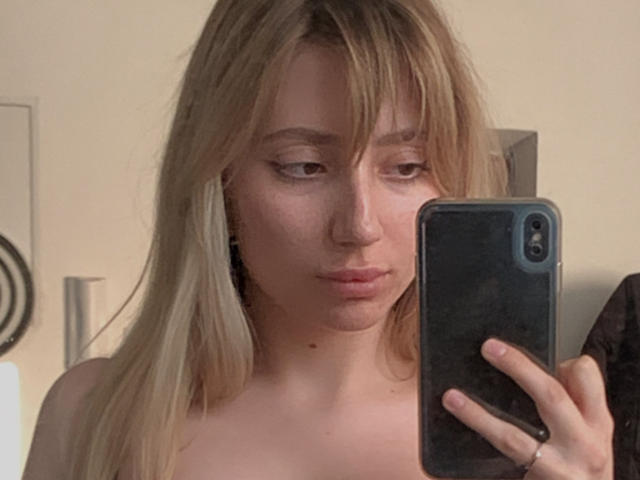 NicoleNovak with blonde hair, holding a black phone, taking a mirror selfie, showing her bare shoulders, pouty lips, and subtle eyeliner. Her tits are partially visible.