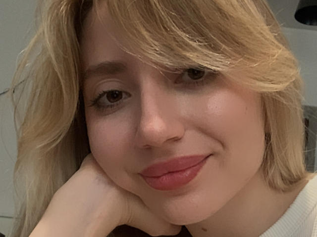 NicoleNovak has blonde hair, pink lips, and a slight smile. She's resting her chin on her hand, looking cute with soft brown eyes and a fair skin tone.