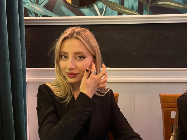 NicoleNovak, blonde, wearing black blazer, holding phone to ear, slightly smiling, black nail polish, blue-green wallpaper in background, wooden chair, dark green curtain on left.