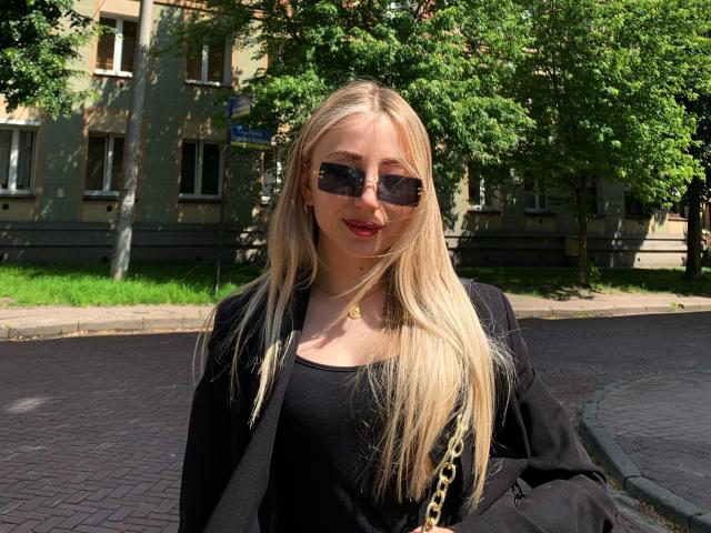 NicoleNovak with long blonde hair, wearing black sunglasses, red lipstick, black top, black jacket, gold necklace, black bag with gold chain, standing on a sunny cobblestone street.