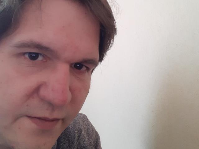 NectarineFad has a pale, slightly red nose with small blemishes, brown hair, and a subtle smile. His eyes are dark and slightly squinted. He's wearing a brown shirt.