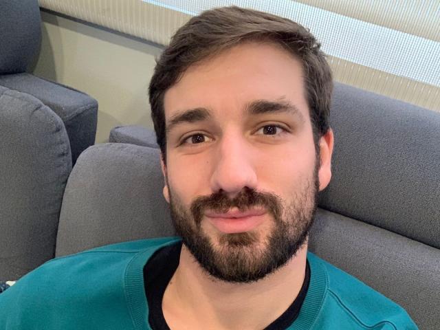NaughtyGuyX has a light skin tone, brown eyes, short brown hair, beard, and is wearing a teal sweater with a black shirt underneath, smiling at the camera.