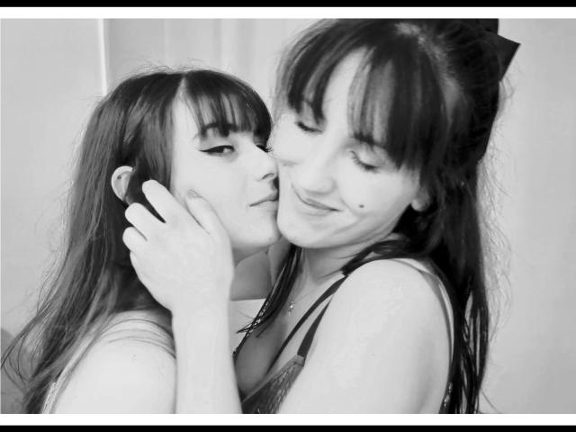 TuttiFloyd69 with long dark hair, holding another TuttiFloyd69's face, smiling, showing a small beauty mark on the cheek, in black-and-white, intimate, soft, and slightly playful.