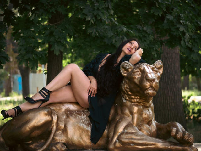 GinaONeon with long black hair, lying on a gold lion statue, wearing a black dress, high heels, and showing her ass, in a tree-filled outdoor setting.