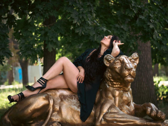 GinaONeon lounges on a bronze lion statue, legs crossed, black dress, high heels, long dark hair, eyes closed, hand in hair, golden sunlight, leafy background, sexy.