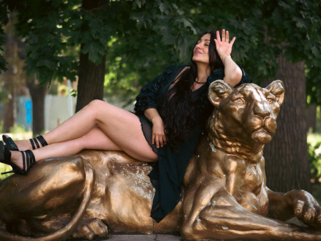 GinaONeon with long black hair, black dress, and black heels, lounging on a bronze lion statue, her hand raised, legs crossed, showing her ass, in a park.