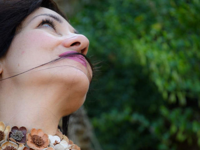 GinaONeon's face is up, showing her nose and lips with dark hair and purple lipstick, wearing floral necklaces, looking upwards with a slight smile.