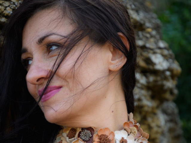 GinaONeon has dark hair, pink lipstick, and a flower necklace. Her face is slightly turned, with a small, knowing smile. Her skin is light, and she's looking to the side.