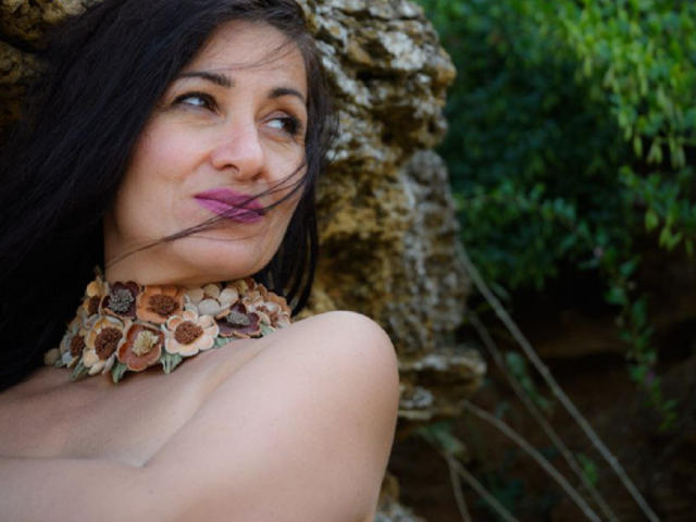 GinaONeon with dark hair, purple lipstick, and floral necklace, arms crossed, leaning against rock, greenery in background, looking to the side with a slight smile.