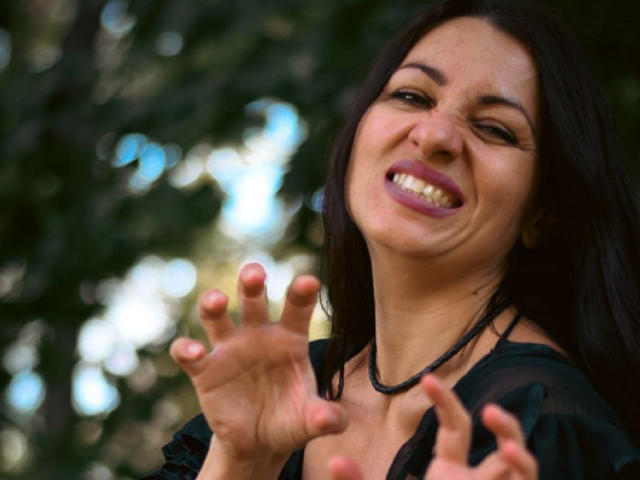 GinaONeon with dark hair, smiling, showing teeth, wearing purple lipstick, black necklace, and black top, hands raised, fingers spread, looking happy and playful.