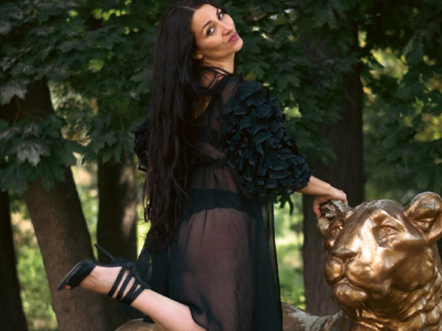 GinaONeon with long black hair, sheer black dress, ruffled sleeves, black heels, and her ass raised, leaning on a gold lion statue in a forest.