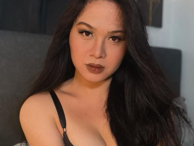 RichCarla with long black hair, brown eyes, and dark lipstick, wearing a black strap bra, showing her tits, looking directly at the camera.