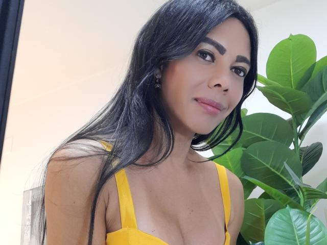 DaianaModeX with long black hair, yellow top, showing her tits, green plant in background, smiling, brown eyes, pink lipstick, earrings, light skin, and a cute face.