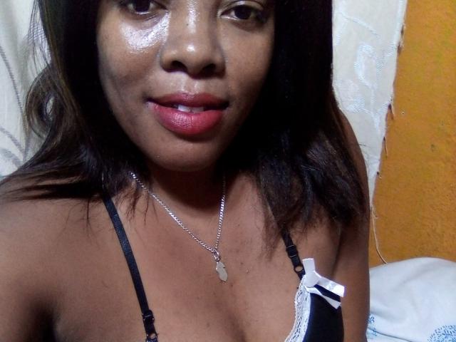 BrownieBabe with shiny brown skin, red lipstick, black lace bra, white bow, silver necklace, and medium-sized tits, looking at camera with slight smile.