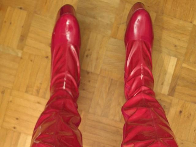 Bootsica-hot's shiny red boots with crisscross laces are standing on a wooden parquet floor, showing the soles and the upper part of their calves.
