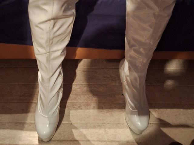 Bootsica-hot's white, shiny, knee-high, criss-cross, leather boots with pointed toes and small heels are standing on a wooden floor, casting subtle shadows.