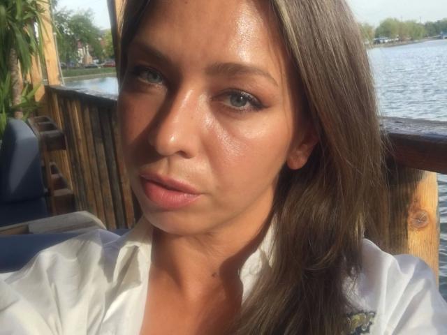 XEvelynxx-hot has shiny, sweaty skin, brown eyes, and brown hair. She's wearing a white shirt, looking serious with a slight pout, and is outdoors by a waterway.