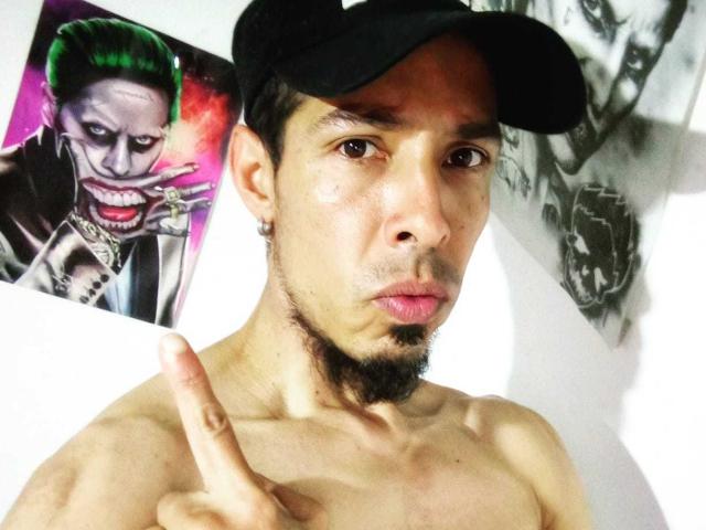 Shirtless JeikkoHot with a black cap, pointing with his middle finger, green-haired Joker poster behind him, and a goatee. His face is serious, brown eyes focused.