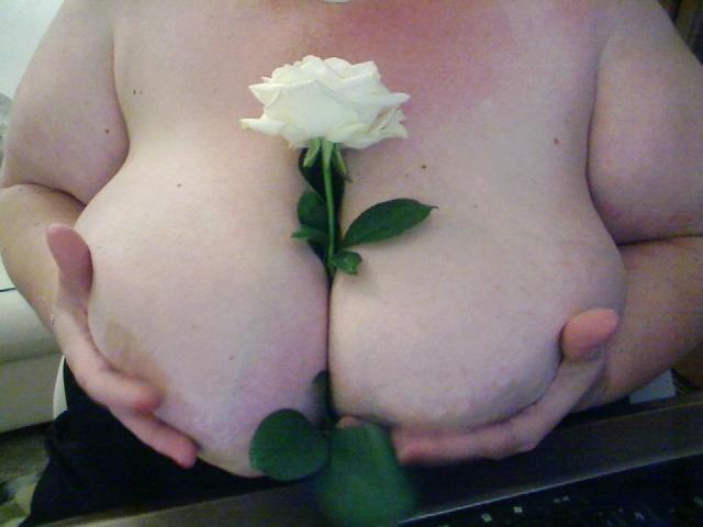 SECRET-CAM's huge, pale tits are held by her hands with a white rose tucked between them. She's got red nails and a silver bracelet, sitting in front of a laptop.