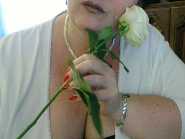 SECRET-CAM with white rose, holding it up to her chest, wearing a white robe and silver necklace, showing her tits and a green leaf tattoo below her boobs.