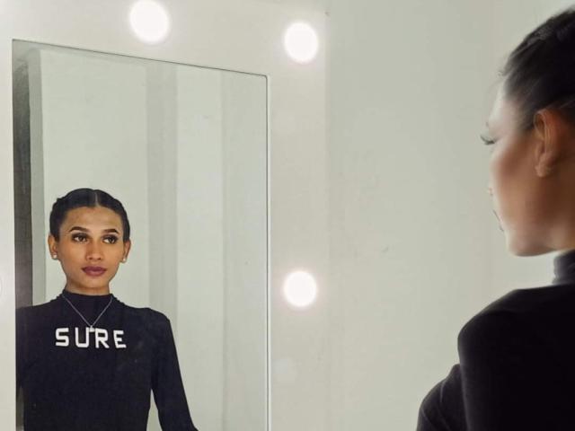 SperanzaFoxy stands in front of a mirror, wearing a black long-sleeve top, with her hair in a tight bun, looking at her reflection, with three bright lights.