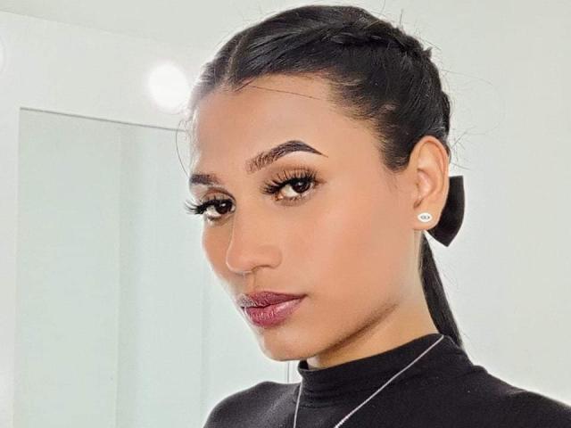 SperanzaFoxy has dark hair in a braid, brown eyes, thick eyebrows, long lashes, pink lipstick, black top, and a black bow in her hair with diamond earrings.