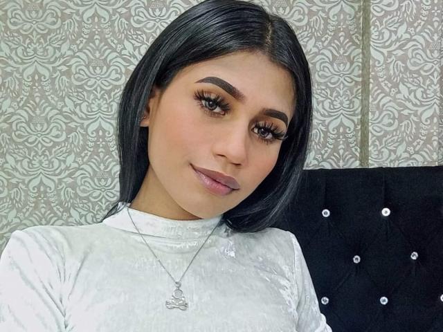 SperanzaFoxy has medium-length black hair, brown eyes, and full lips with pink lipstick. She's wearing a white velvet top and a silver necklace with a small pendant.