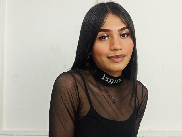SperanzaFoxy with long black hair, brown eyes, and dark lipstick, wearing a sheer black top, black choker with "Jenma" text, and a black necklace.