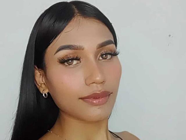 SperanzaFoxy has tan skin, long black hair, thick eyebrows, long eyelashes, brown eyes, and glossy pink lips. She wears small hoop earrings.