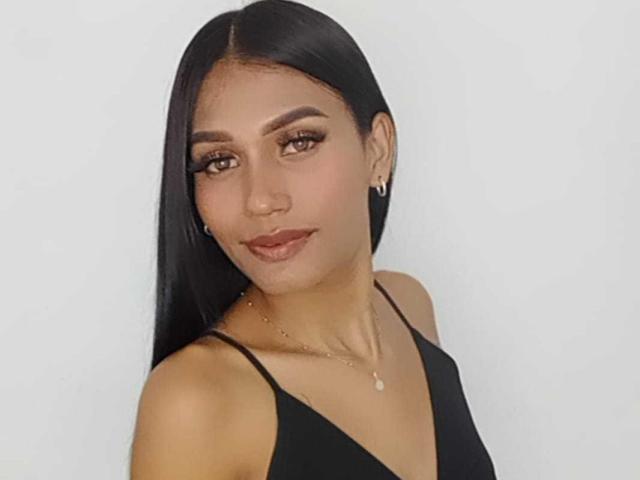 SperanzaFoxy with long black hair, brown eyes, and dark lipstick, wearing a black halter top, showing her bare shoulders, and a small pendant necklace.