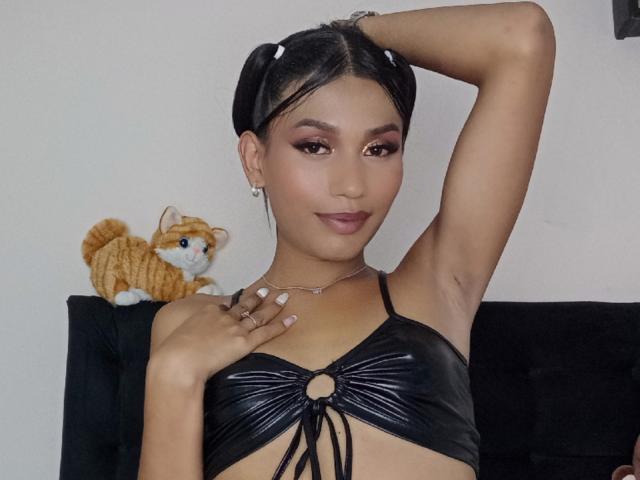 SperanzaFoxy with dark hair in pigtails, black shiny bra, raised arm, cat toy on black couch, small earrings, smiling, cute, brown eyes, tan skin, sexy.