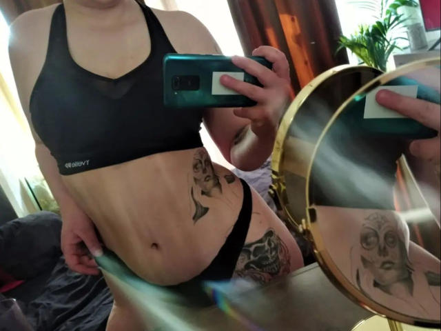 Raubmiez-hot, vestindo um sutiã e calcinha esportivos pretos, mostrando tatuagens na cintura e na coxa, segurando o celular em uma selfie no espelho. Seus seios são pequenos, e sua bunda não está visível..