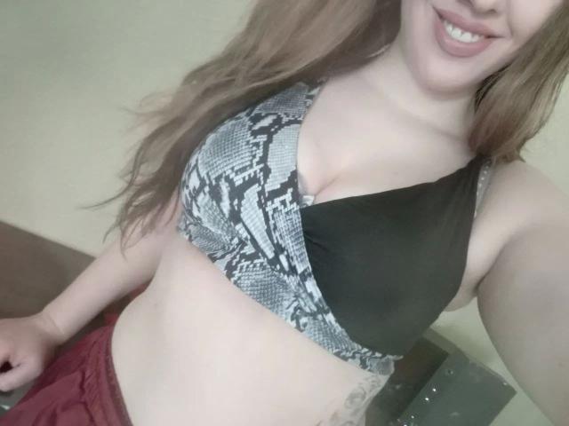 Raubmiez-hot mit welligem, braunem Haar, lächelnd, trägt einen schwarzen und grauen Sport-BH mit Muster, der ihre Brüste zeigt, und rote Hosen, mit einem Tattoo an der Seite..