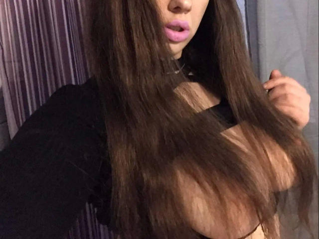Raubmiez-hot, com longos cabelos castanhos, lábios rosados, blusa preta que expõe seios grandes e nus, segurando o cabelo, usando um colar preto, em um quarto com pouca luz..