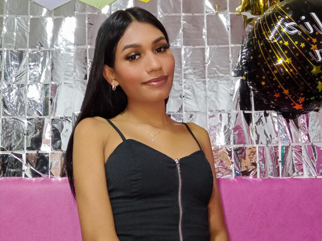 SperanzaFoxy, dark-haired girl, black zip-up dress, small tits, pink lipstick, earrings, silver background, smiling, balloon on right, pink cushion.