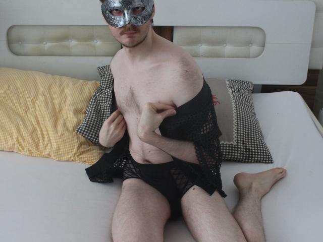 Sweetbody23-hot wearing a silver masquerade mask, black mesh robe, and black underwear, sitting on a bed with yellow and checkered pillows, exposing a bare, hairy chest.