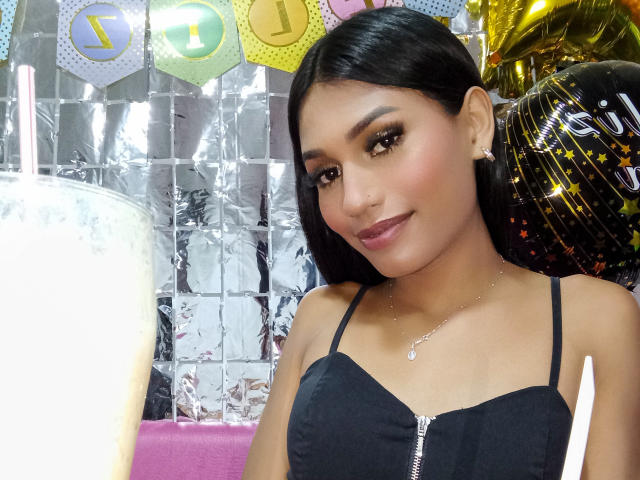 SperanzaFoxy with long black hair, brown eyes, and dark makeup, wearing a black zip-up top, smiling at a drink with a straw, surrounded by balloons and festive decorations.