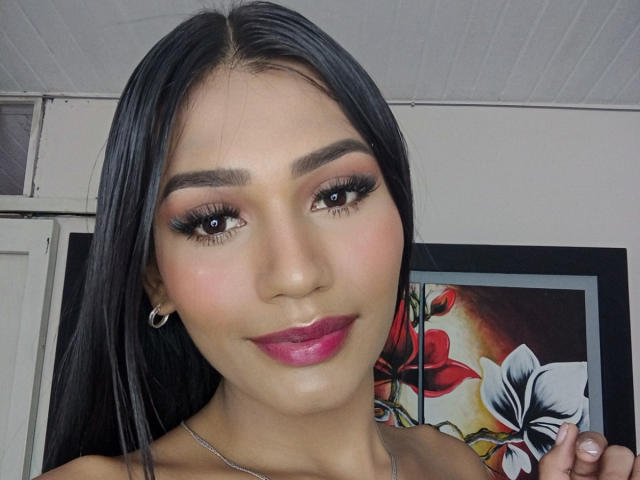 SperanzaFoxy with long black hair, brown eyes, pink lipstick, and black eyeliner, smiling at the camera. She's wearing small hoop earrings and a silver necklace.