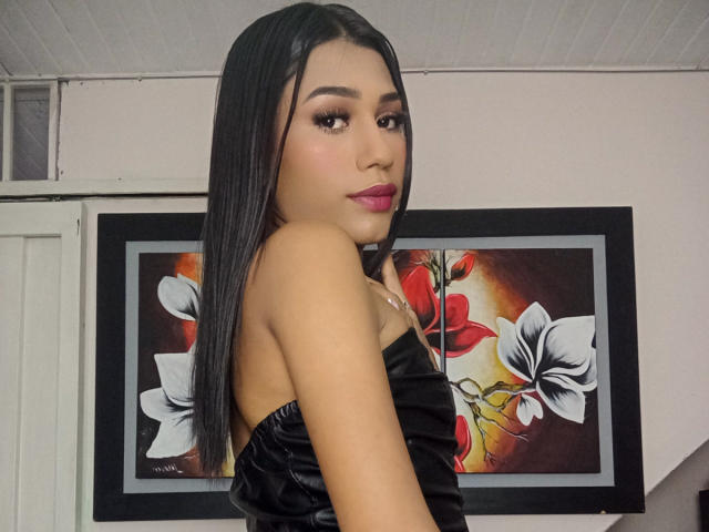 SperanzaFoxy with long black hair, looking over her shoulder, wearing a black strapless top, showing her small ass, and red lipstick, in a room with floral art.