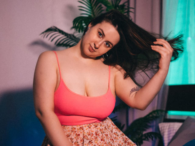ViaLattea with medium tits in a pink tank top, holding her brown hair, wearing floral pants, has a tattoo on her right arm, and a cute smile.