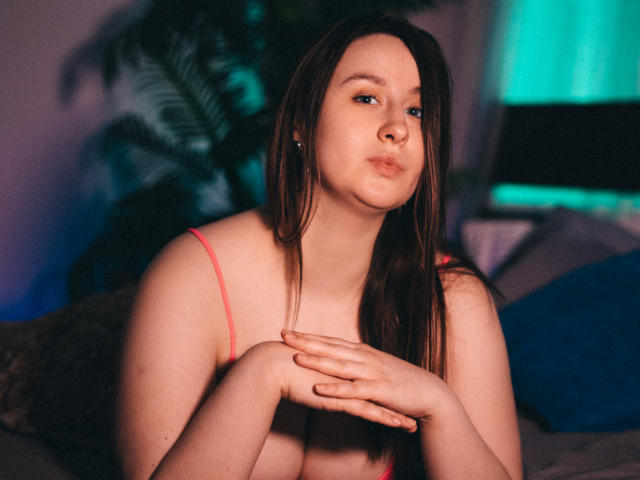 ViaLattea with pouty lips, brown hair, and pink tank top, hands crossed, looking slightly to the side, in a dimly lit room with green and blue lights.