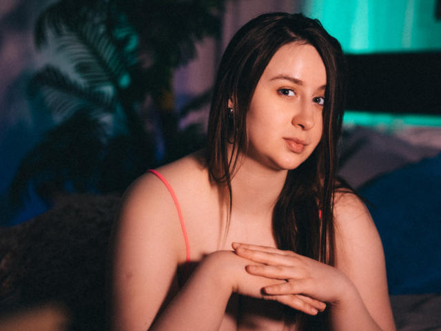 ViaLattea with wet brown hair, pink bra, and crossed arms, pouting slightly, has soft skin and small tits, in a dimly lit room with green and blue lights.