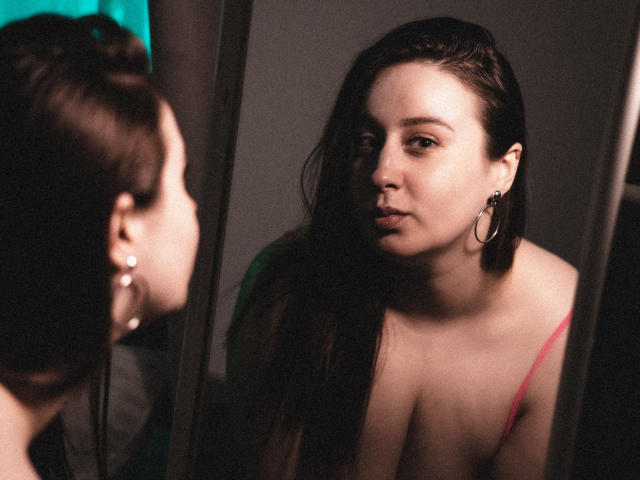 ViaLattea with long brown hair, wearing a pink strap, looking in a mirror, pouty face, hoop earrings, soft light, reflection shows her tits, dark background.