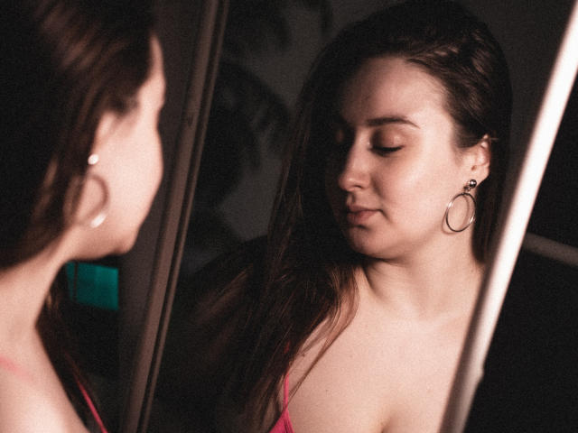 ViaLattea with long brown hair, wearing hoop earrings and pink top, looking at herself in a mirror with a serious expression, showing her tits in the reflection.