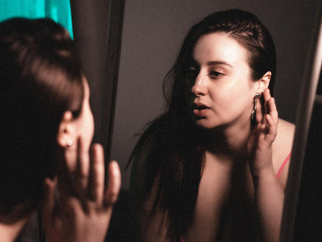 ViaLattea with wet, dark brown hair, looking in a mirror, touching her ear with a hand, wearing a pink top, showing a slight, serious expression, soft lighting.