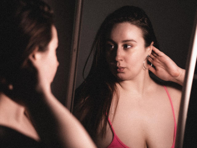 ViaLattea with long brown hair, wearing a pink spaghetti strap top, stares into a mirror, touching her ear, showing her tits and a serious expression. Dark, moody lighting.