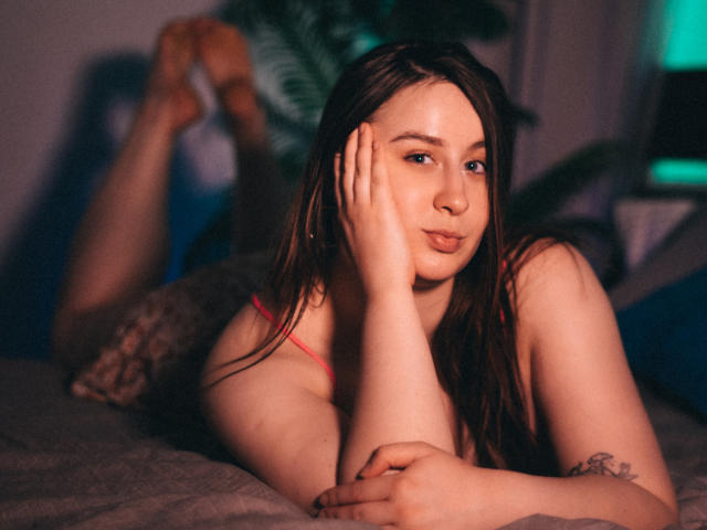 ViaLattea with long brown hair, laying on a bed, wearing a red spaghetti strap top, hand on cheek, looking at camera, legs up, soft light, slight smile, tattoo on arm.