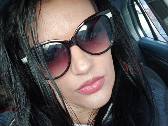 KissYouKissMe has glossy pink lips, dark sunglasses with pink lenses, black straight hair, and a nose piercing, sitting in a car, wearing a black jacket.