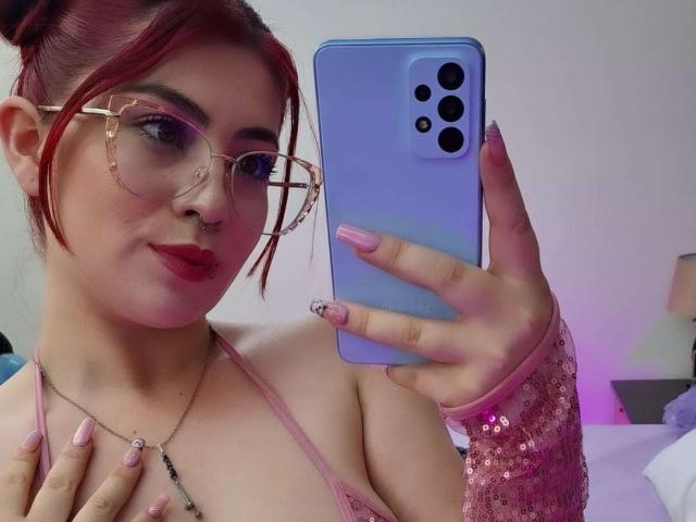IsabellaRobertz, com cabelo ruivo, óculos, batom rosa, unhas rosa, capa de celular rosa, com lantejoulas rosa no braço, segurando o celular, tirando uma selfie, mostrando seios pequenos, em uma selfie no espelho..