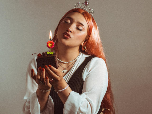 IsabellaClown with red hair, wearing a tiara, holding a candle-topped chocolate cake, eyes closed, red lipstick, white shirt, black vest, jewelry, red nails, slightly pouty expression.